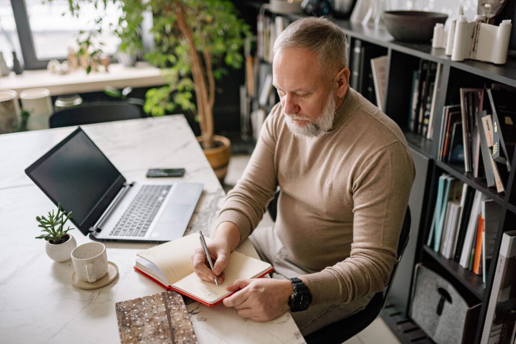 Senior man is working at a desk with a laptop and a journal